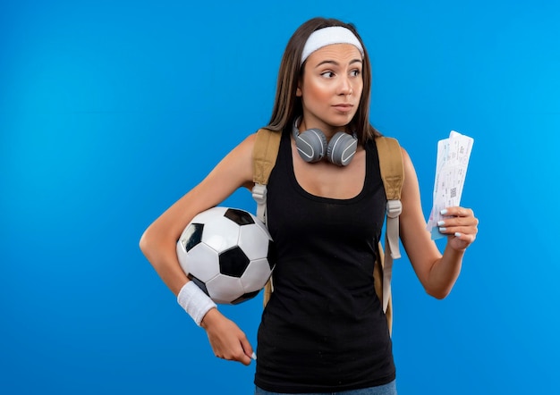 Onder de indruk jong mooi sportief meisje met hoofdband en polsbandje en rugtas met koptelefoon op nek met vliegtickets met voetbal geïsoleerd op blauwe muur met kopieerruimte