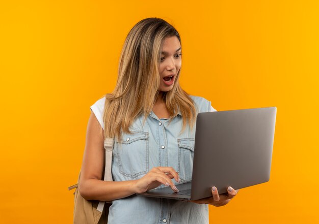 Onder de indruk jong mooi studentenmeisje die achterzak dragen die en laptop gebruikt die op oranje wordt geïsoleerdk