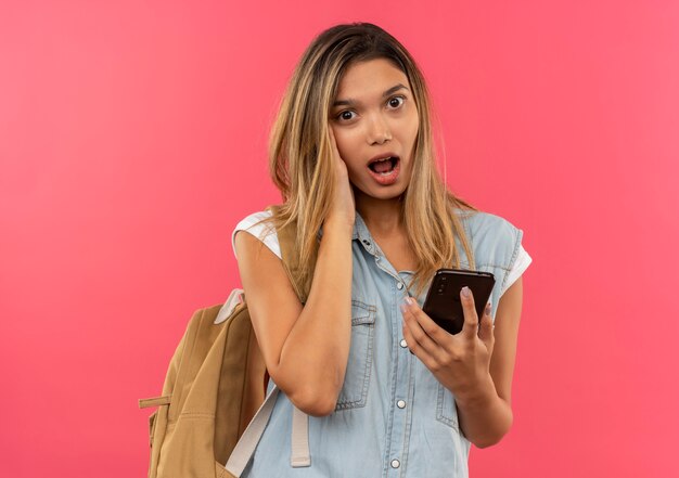 Onder de indruk jong mooi studentenmeisje die achterzak dragen die mobiele telefoon met hand op gezicht houdt dat op roze wordt geïsoleerd