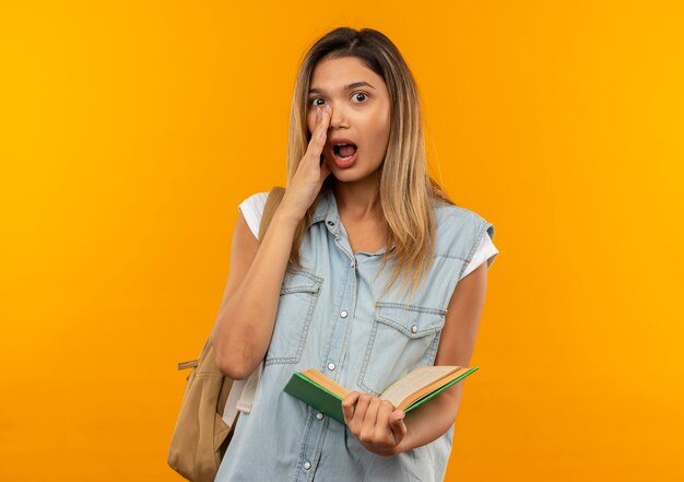 Onder de indruk jong mooi studentenmeisje die achterzak dragen die open boek houden dat hand dichtbij mond zet die aan voorzijde fluistert die op oranje wordt geïsoleerd