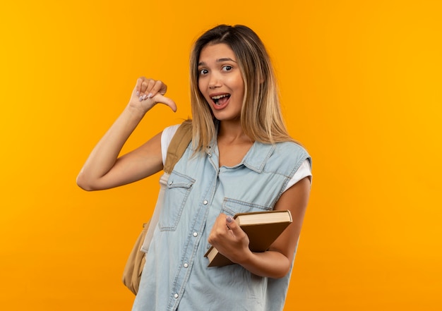 Onder de indruk jong mooi studentenmeisje die achterzak dragen die open boek houdt die op zichzelf richt dat op oranje wordt geïsoleerd