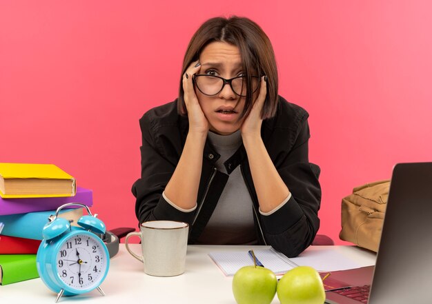 Onder de indruk jong studentenmeisje die glazen dragen die aan bureau zitten dat huiswerk doet die handen op tempels zetten die op roze worden geïsoleerd