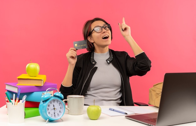 Onder de indruk jong studentenmeisje die glazen dragen die bij de creditcard van de bureauholding zitten die vinger opheft die op roze wordt geïsoleerd