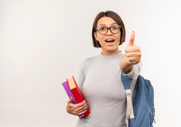 Onder de indruk jong studentenmeisje die glazen en de boeken van de achterzakholding dragen die duim tonen die omhoog op wit wordt geïsoleerd