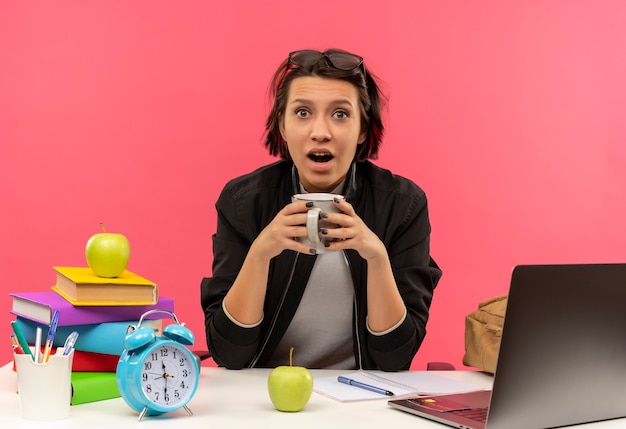 Onder de indruk jong studentenmeisje die glazen op hoofd dragen die kop van koffie houden die aan bureau huiswerk doen dat op roze wordt geïsoleerd