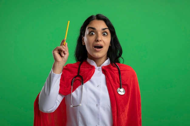 Onder de indruk jong superheromeisje die medisch kleed met stethoscoop dragen dat potlood opheft dat op groen wordt geïsoleerd