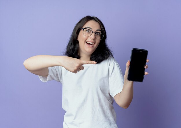 Onder de indruk jong vrij Kaukasisch meisje dat een bril draagt die en op mobiele telefoon toont richt die op purpere achtergrond wordt geïsoleerd