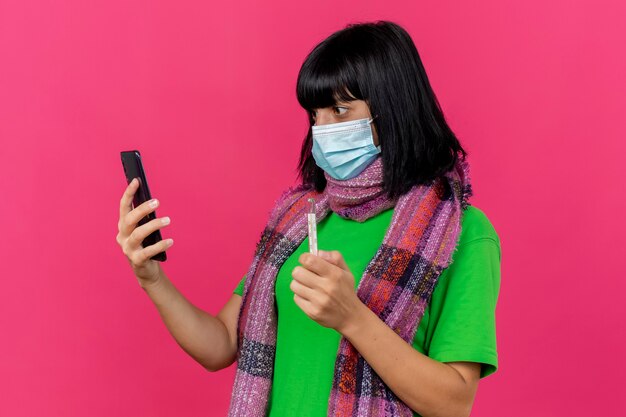 Onder de indruk jong ziek Kaukasisch meisje die masker en sjaal dragen die thermometer en mobiele telefoon houden die telefoon bekijken die op karmozijnrode achtergrond met exemplaarruimte wordt geïsoleerd