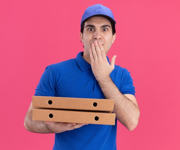 Onder de indruk jonge bezorger in blauw uniform en pet met pizzapakketten die hand op de mond zetten en naar de voorkant kijken geïsoleerd op roze muur