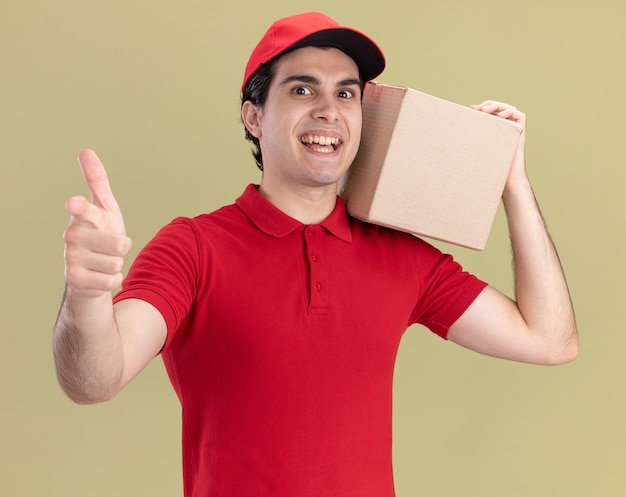 Onder de indruk jonge bezorger in rood uniform en pet met kartonnen doos op schouder kijkend en wijzend naar voren geïsoleerd op olijfgroene muur