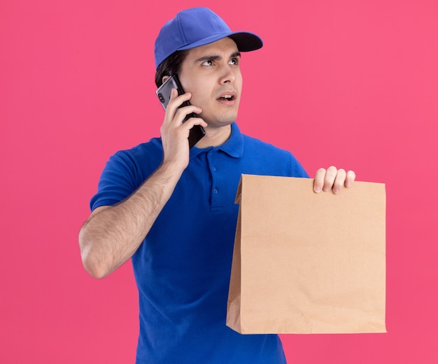 Onder de indruk jonge blanke bezorger in blauw uniform en pet met papieren pakket pratend aan de telefoon en geïsoleerd op roze muur