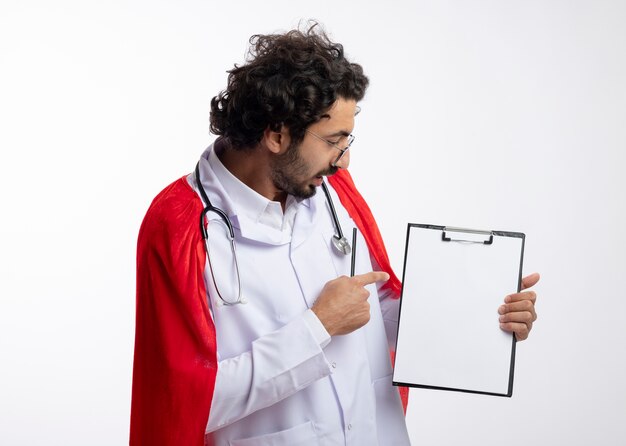 Onder de indruk jonge blanke man in optische bril dragen arts uniform met rode mantel en met een stethoscoop om de nek kijkt en wijst naar het klembord met potlood geïsoleerd op een witte muur