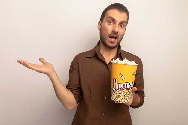 Onder de indruk jonge blanke man met emmer popcorn met lege hand kijken naar camera geïsoleerd op een witte achtergrond met kopie ruimte