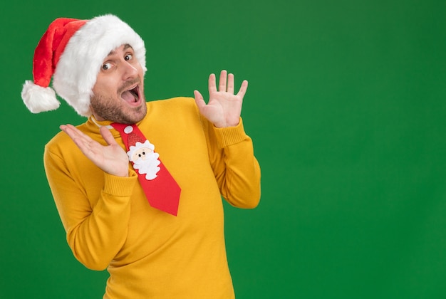 Onder de indruk jonge blanke man met kerstmuts en stropdas kijken camera houden handen in de lucht geïsoleerd op een groene achtergrond