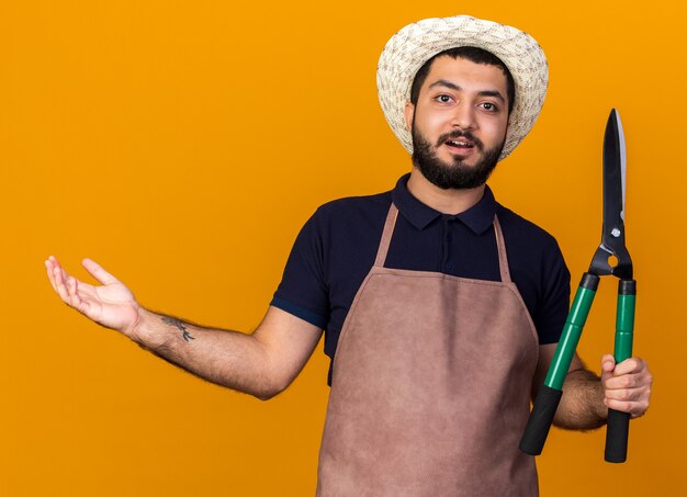 onder de indruk jonge blanke mannelijke tuinman tuinieren hoed bedrijf tuinieren schaar en houden hand open geïsoleerd op oranje muur met kopie ruimte