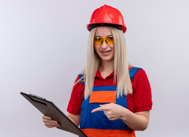 Onder de indruk jonge blonde ingenieur bouwer meisje in uniform in tandheelkundige accolades dragen veiligheidsbril klembord houden en erop gericht op geïsoleerde witte ruimte