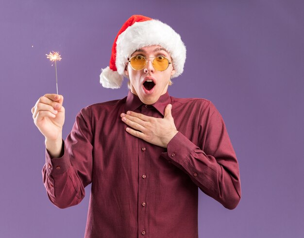 Onder de indruk jonge blonde man met kerstmuts en bril met vakantie sterretje kijken camera houden hand op borst geïsoleerd op paarse achtergrond