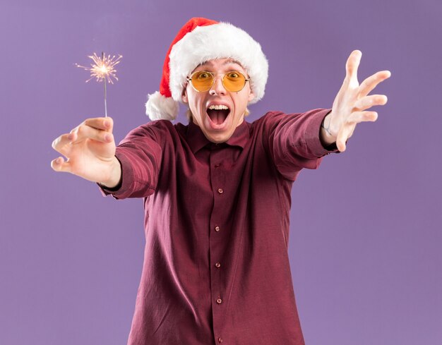 Onder de indruk jonge blonde man met kerstmuts en bril vakantie sterretje uitrekken en hand doen welkom gebaar geïsoleerd op paarse muur