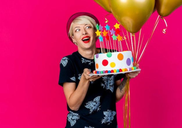 Onder de indruk jonge blonde partijvrouw die partijhoed draagt die ballons en verjaardagstaart met sterren houdt die cake bekijken die op karmozijnrode muur wordt geïsoleerd