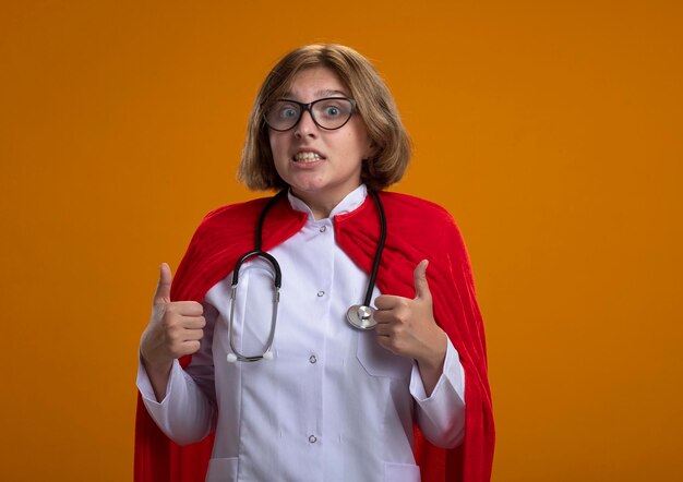 Onder de indruk jonge blonde superheld vrouw in rode cape arts uniform en bril met stethoscoop kijken voorzijde tonen duimen omhoog geïsoleerd op muur met kopie ruimte