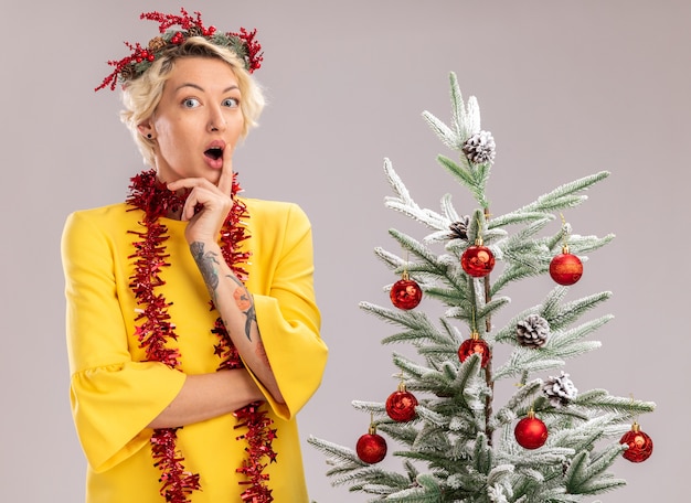 Onder de indruk jonge blonde vrouw die de hoofdkrans van Kerstmis en een klatergoudslinger om de hals draagt die zich dichtbij versierde kerstboom bevindt die op zoek raakt lippen geïsoleerd op witte muur