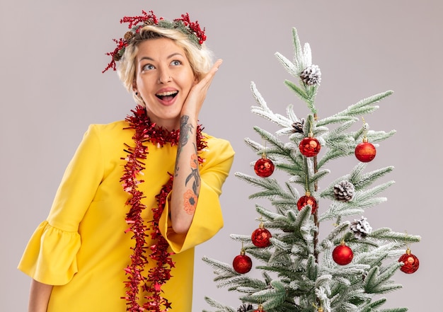 Onder de indruk jonge blonde vrouw die de hoofdkrans van Kerstmis en een klatergoudslinger om de hals draagt en zich dichtbij versierde kerstboom bevindt die gezicht aanraken kijkt naar kant geïsoleerd op witte muur