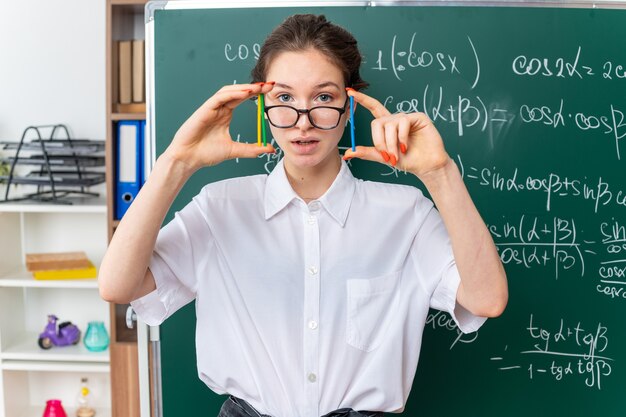 Onder de indruk jonge blonde vrouwelijke wiskundeleraar met een bril die voor een schoolbord staat met telstokken die naar de camera in de klas kijken
