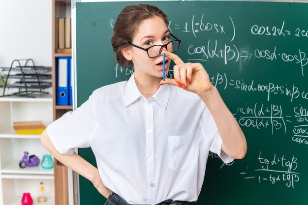 Onder de indruk jonge blonde vrouwelijke wiskundeleraar met een bril die voor het bord staat en de hand op de taille houdt met de telstok in de klas