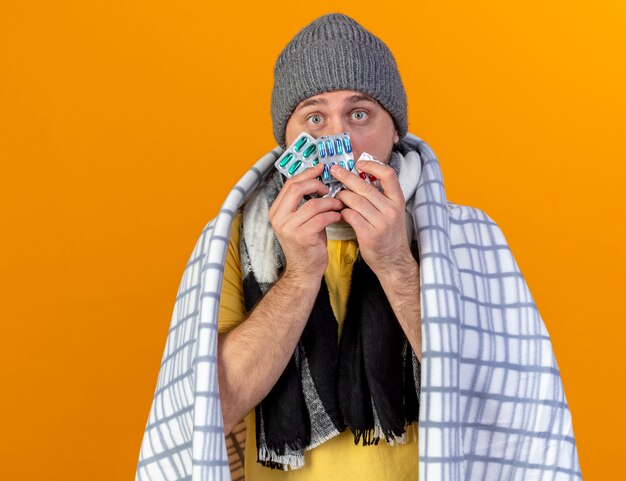 Gratis foto onder de indruk jonge blonde zieke slavische man met winter muts en sjaal gewikkeld in plaid houdt verpakkingen van medische pillen geïsoleerd op oranje muur met kopie ruimte
