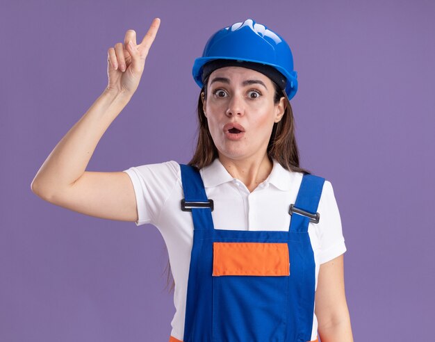 Onder de indruk jonge bouwersvrouw in uniforme punten omhoog geïsoleerd op paarse muur