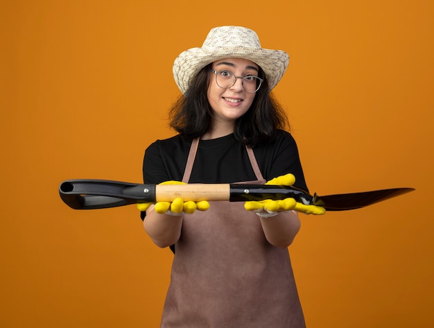 Onder de indruk jonge brunette vrouwelijke tuinman in optische bril en uniform dragen tuinieren hoed en handschoenen houdt schop geïsoleerd op oranje muur