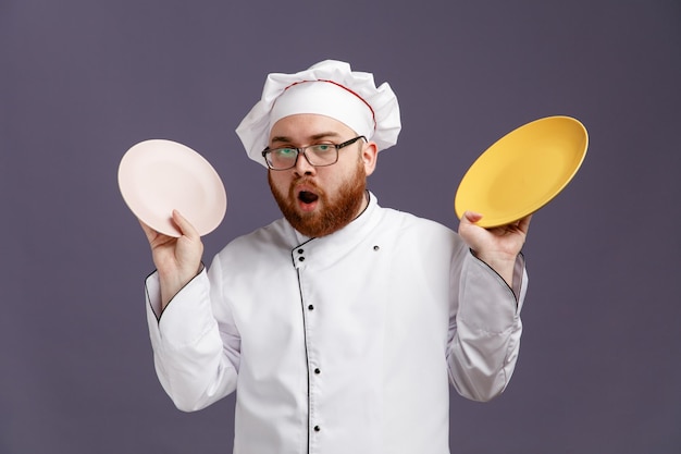 Onder de indruk jonge chef-kok met een uniforme bril en een pet die naar de camera kijkt die lege borden opheft en ze laat zien geïsoleerd op een paarse achtergrond
