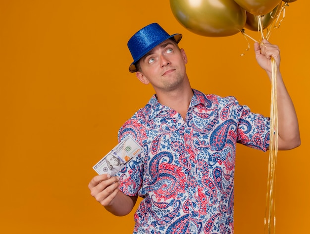 Onder de indruk jonge feest man met blauwe hoed met ballonnen met contant geld geïsoleerd op oranje