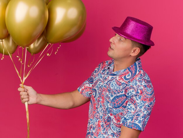 Onder de indruk jonge feestmens die een roze hoed draagt en naar ballonnen kijkt die op roze worden geïsoleerd