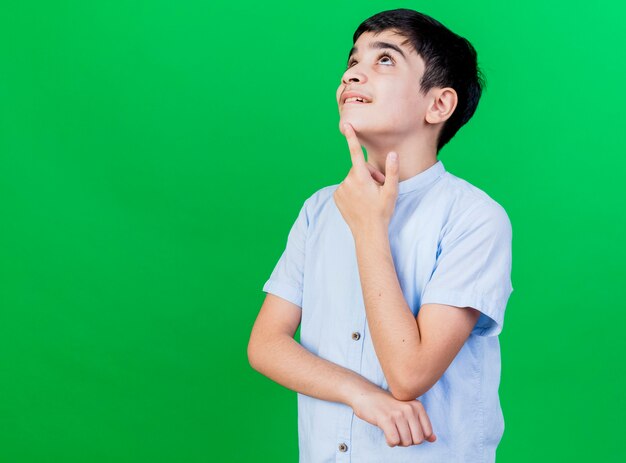 Onder de indruk jonge jongen die vinger op kin zet die omhoog geïsoleerd op groene muur kijken