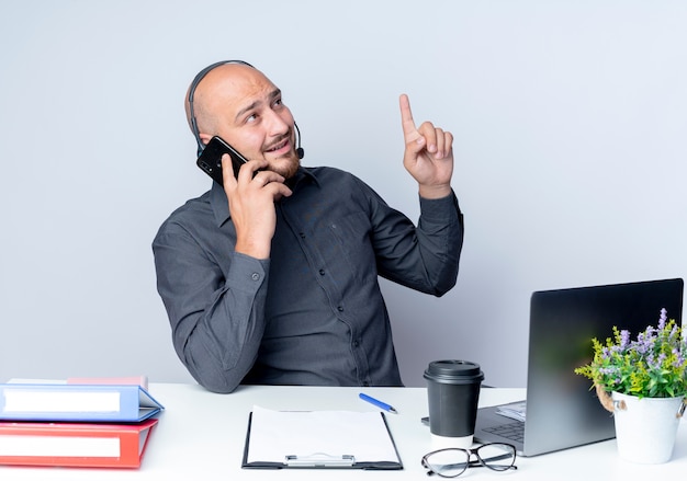 Onder de indruk jonge kale call center man met hoofdtelefoon zit aan bureau met uitrustingsstukken praten over telefoon verhogen vinger en kijken naar kant geïsoleerd op wit