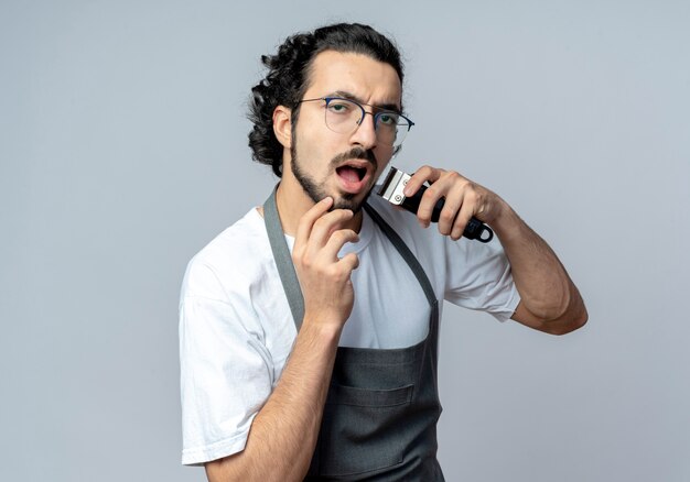 Onder de indruk jonge Kaukasische mannelijke kapper bril en golvende haarband dragen uniform zijn baard trimmen met een tondeuse en aanraken van zijn kin geïsoleerd op een witte achtergrond met kopie ruimte