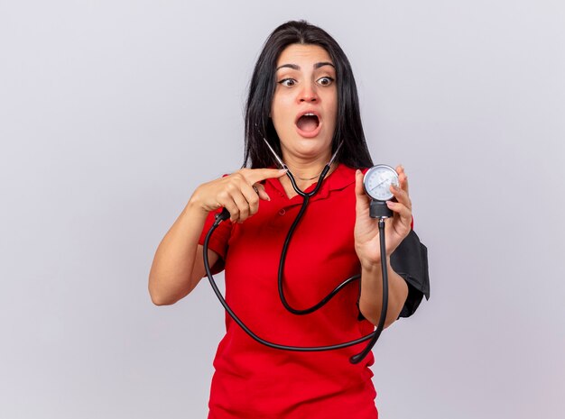 Onder de indruk jonge Kaukasische ziek meisje dragen stethoscoop haar druk meten met bloeddrukmeter kijken en wijzend op het geïsoleerd op een witte achtergrond met kopie ruimte