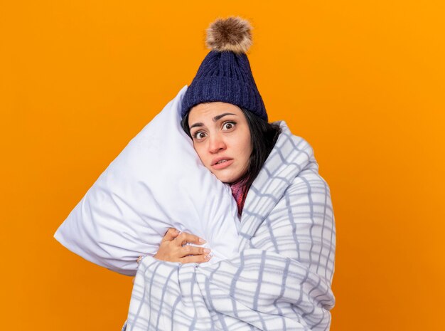 Onder de indruk jonge Kaukasische ziek meisje dragen winter hoed en sjaal verpakt in geruite knuffelen kussen kijken camera geïsoleerd op een oranje achtergrond met kopie ruimte