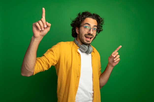 Onder de indruk jonge knappe blanke man met bril met koptelefoon om nek omhoog geïsoleerd op groene muur