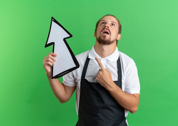 Onder de indruk jonge knappe kapper die eenvormig en het teken van de holdingspijl draagt ?? die omhoog geïsoleerd op groen benadrukt