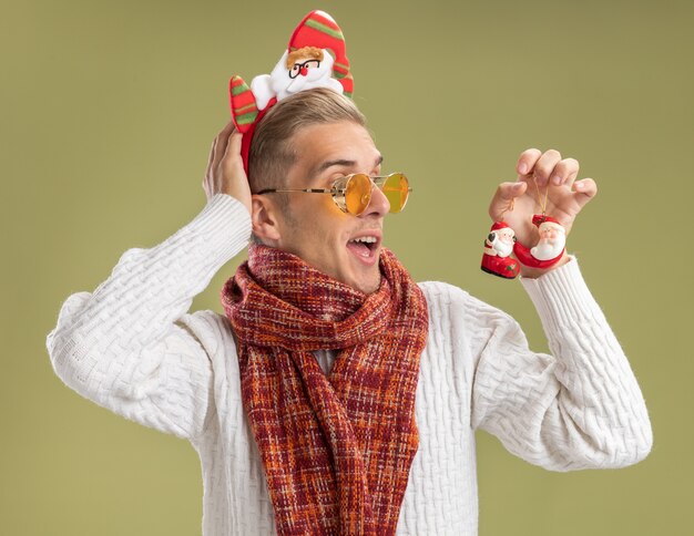 Onder de indruk jonge knappe kerel die de hoofdband en de sjaal van de kerstman draagt en naar de kerstversieringen van de kerstman kijkt, hand achter het hoofd zetten geïsoleerd op olijfgroene muur