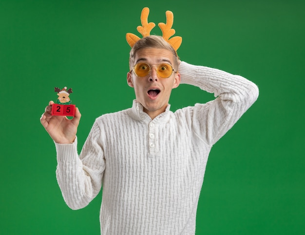 Onder de indruk jonge knappe kerel die rendiergeweien hoofdband met bril draagt die kerstboomstuk speelgoed met datum houdt die hand achter hoofd houdt geïsoleerd op groene muur