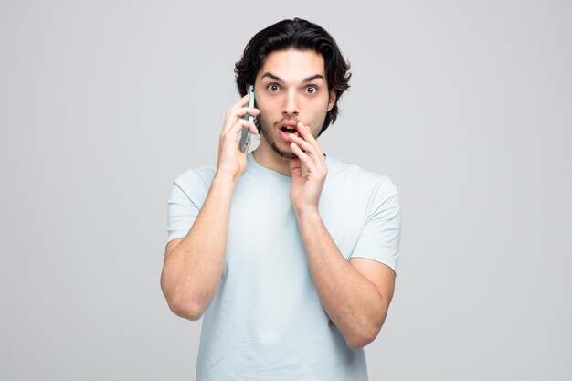 onder de indruk jonge knappe man die hand in de buurt van de mond houdt en naar de camera kijkt terwijl hij aan de telefoon praat op een witte achtergrond