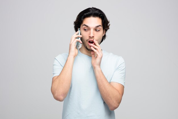 Onder de indruk jonge knappe man die lippen aanraakt en naar de zijkant kijkt terwijl hij aan de telefoon praat fluisterend geïsoleerd op een witte achtergrond