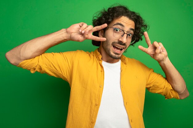 Onder de indruk jonge knappe man met bril doet vredesteken kijken voorzijde geïsoleerd op groene muur