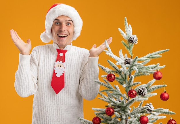 Onder de indruk jonge knappe man met kerstmuts en stropdas van de kerstman staande in de buurt van versierde kerstboom kijken naar camera met lege handen geïsoleerd op een oranje achtergrond