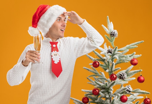 Onder de indruk jonge knappe man met kerstmuts en stropdas van de kerstman staande in de buurt van versierde kerstboom met glas champagne kijken naar kant in afstand geïsoleerd op oranje muur