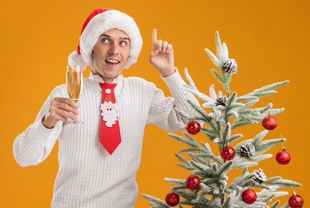 Onder de indruk jonge knappe man met kerstmuts en stropdas van de kerstman staande in de buurt van versierde kerstboom met glas champagne kijken naar kant omhoog geïsoleerd op oranje muur