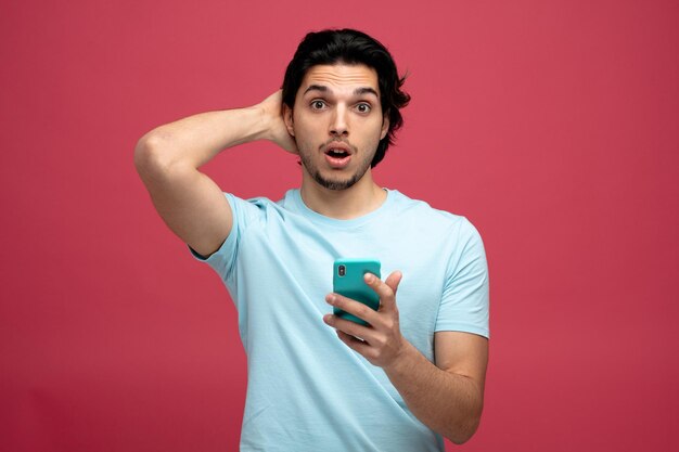 Onder de indruk jonge knappe man met mobiele telefoon die hand achter het hoofd houdt kijkend naar camera geïsoleerd op rode achtergrond